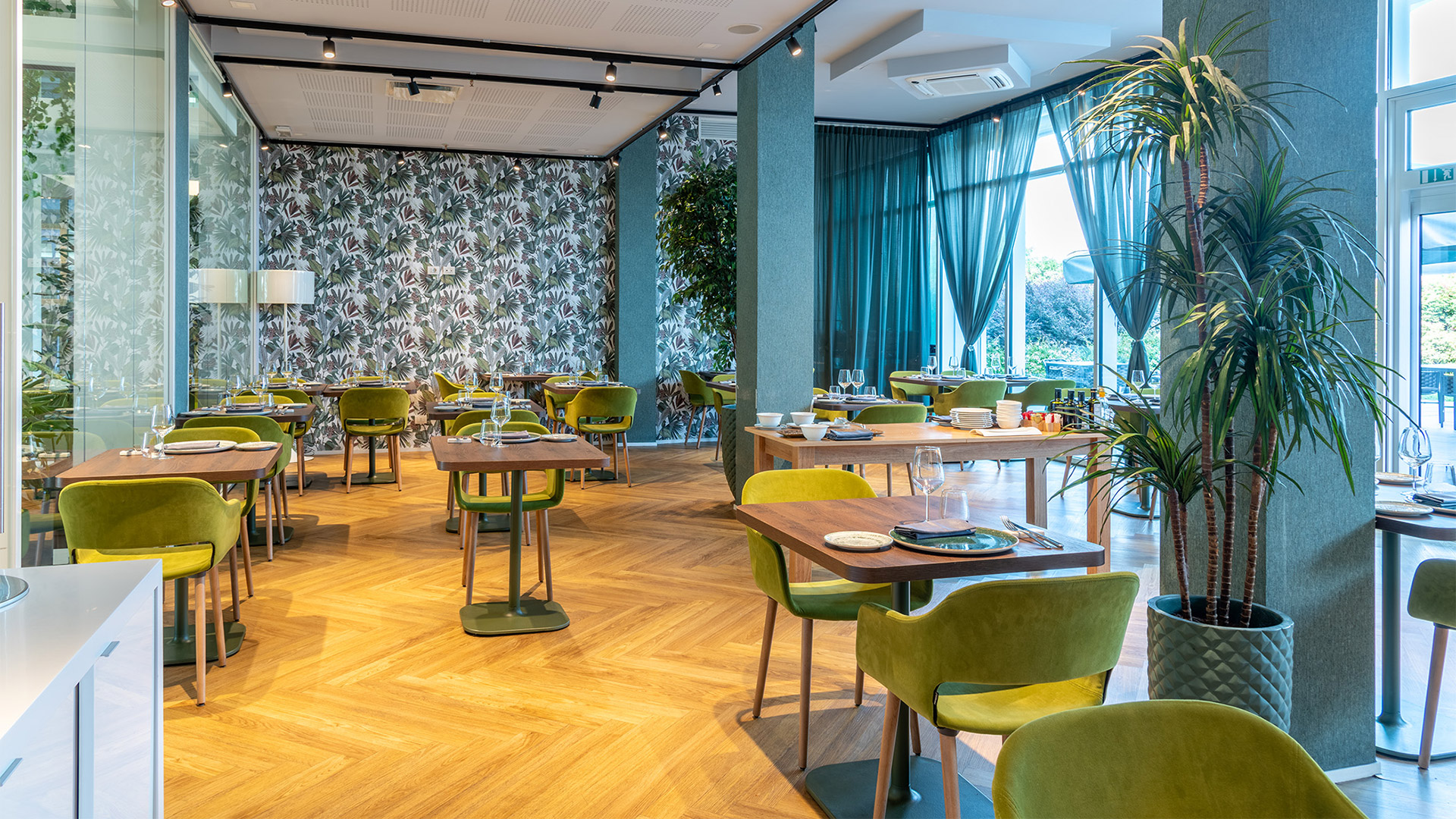 Sala ristorante luminosa con arredi moderni, sedie verde velluto, tavoli in legno e grandi vetrate con tende