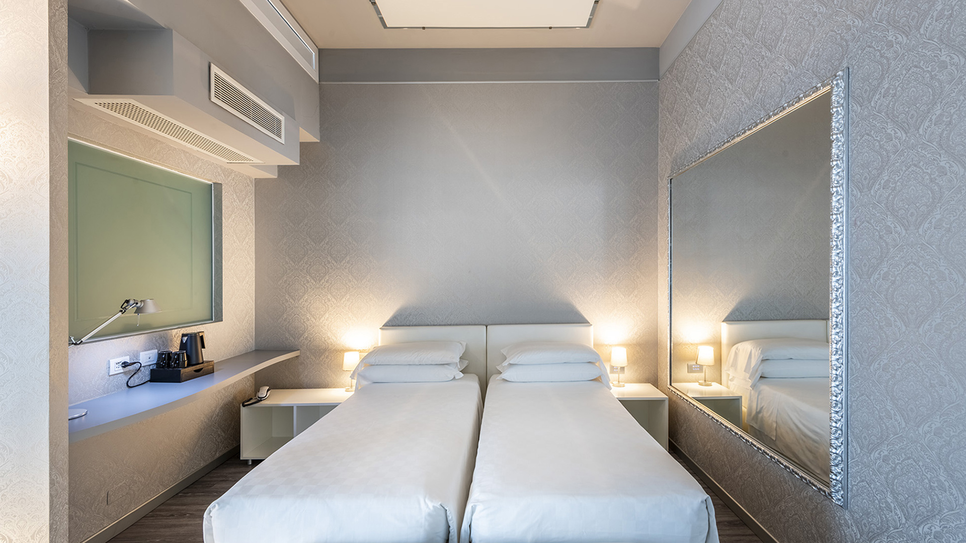 Camera Deluxe con due letti singoli affiancati, comodini con abat-jour, grande specchio a parete con cornice argentata e mensola con bollitore e macchina del caffè