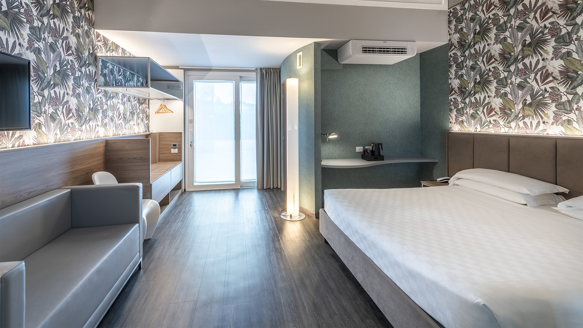 Lussuosa suite Deluxe con luce soffusa, divano grigio, letto, carta da parati con foglie verdi e tende grigie