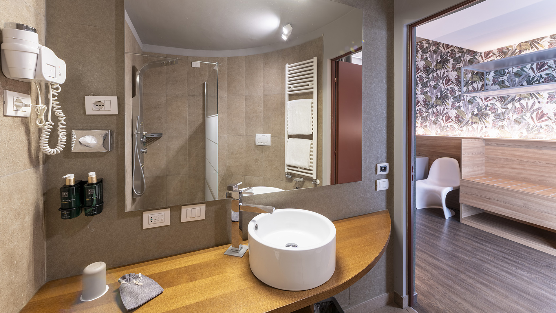 Bagno moderno con lavabo rotondo su piano in legno, ampia specchiera, doccia walk-in e vista sulla camera Deluxe con carta da parati floreale e arredi in legno
