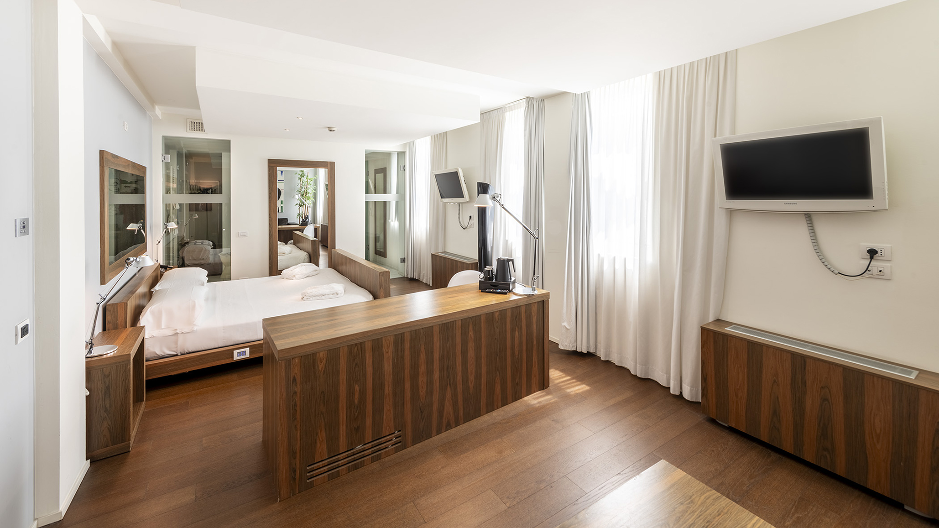 Suite Superior con letto matrimoniale in legno, mobile divisorio con piano scrivania integrato, grande specchio a parete, TV a muro e ampie finestre con tende bianche