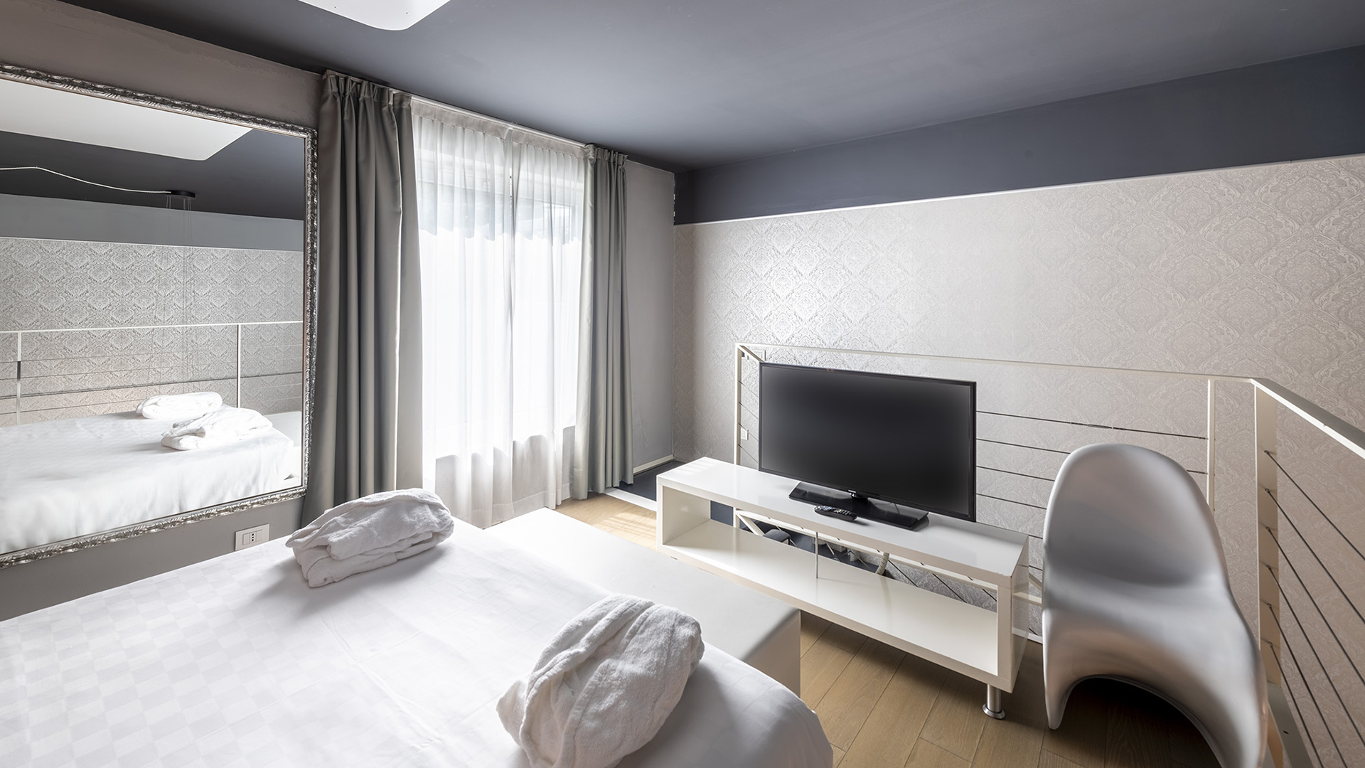 Suite su due livelli con camera matrimoniale al piano superiore, TV su mobile bianco, grande specchio a parete, finestra con tende chiare e sedia di design accanto alla balaustra