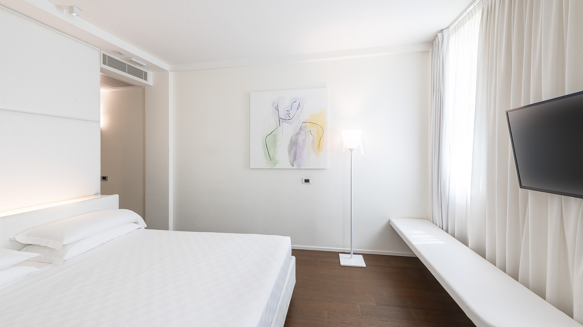 Camera Superior con letto matrimoniale, pareti bianche, quadro astratto, TV a parete, lunga panca sotto la finestra con tende chiare e lampada da terra