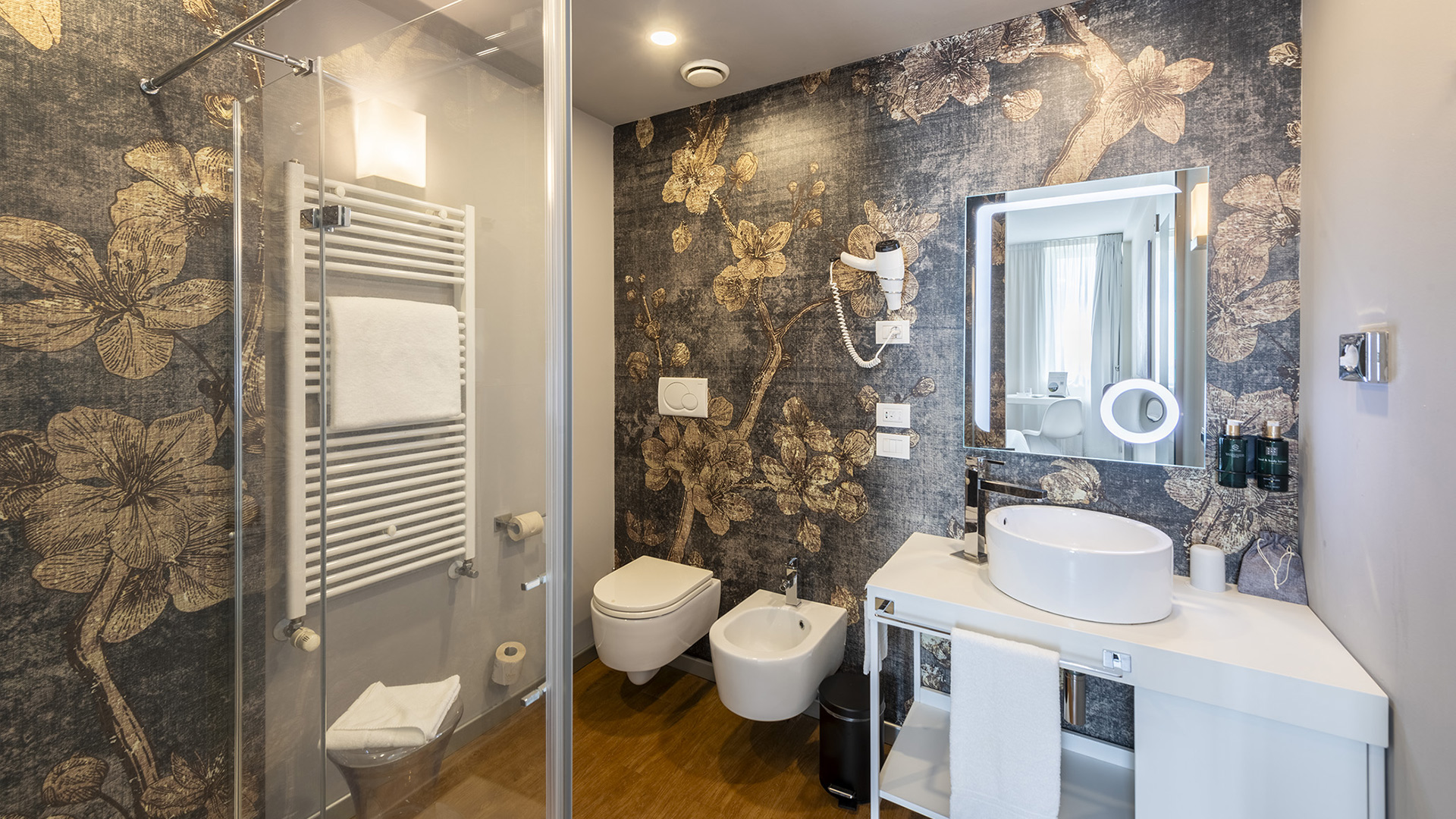 Bagno Superior con doccia in vetro, lavabo da appoggio su piano bianco, specchio retroilluminato e parete decorativa con motivo floreale scuro